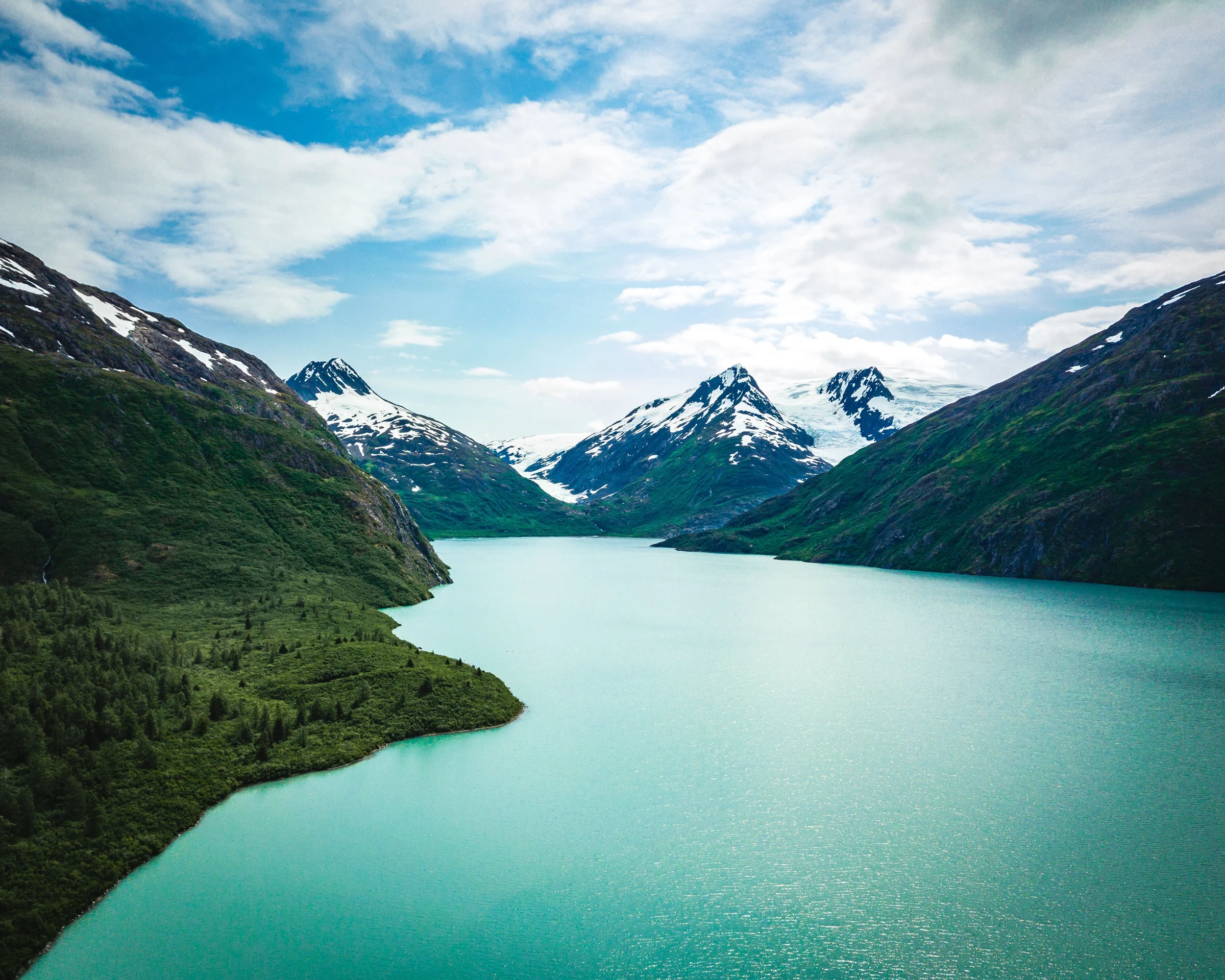 Turquoise lake between green slopes and snow-covered mountains beneath a cloudy blue sky