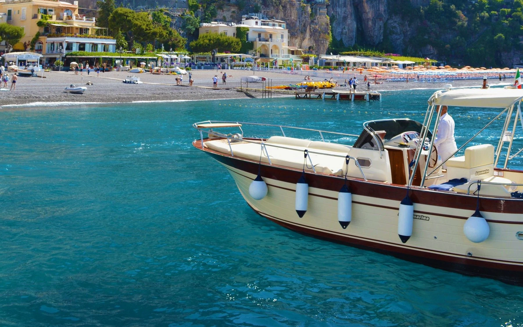 A cream-colored motorboat beside blue water and the Positano waterfront on the Amalfi Coast