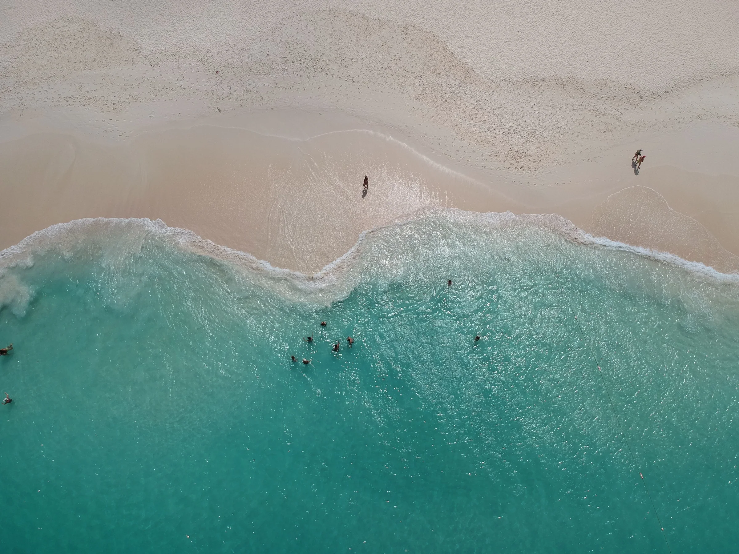 An aerial view of swimmers in turquoise water beside a broad sandy beach