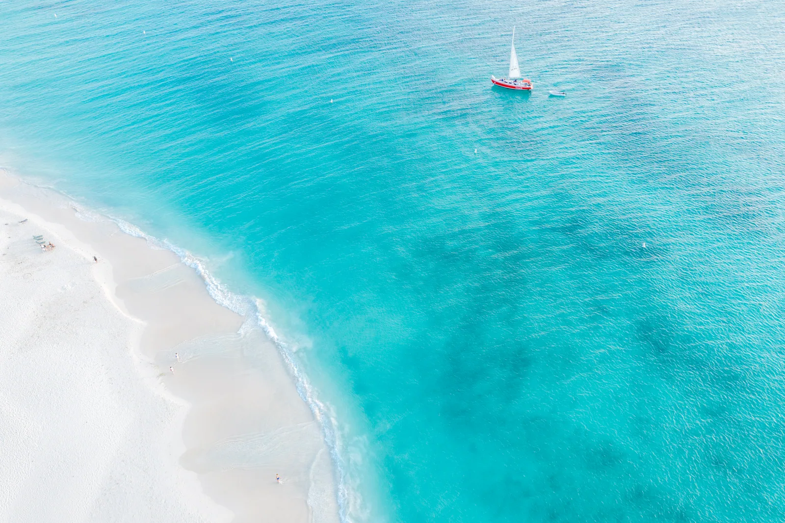 A red-hulled sailboat in turquoise water beside a sandy beach