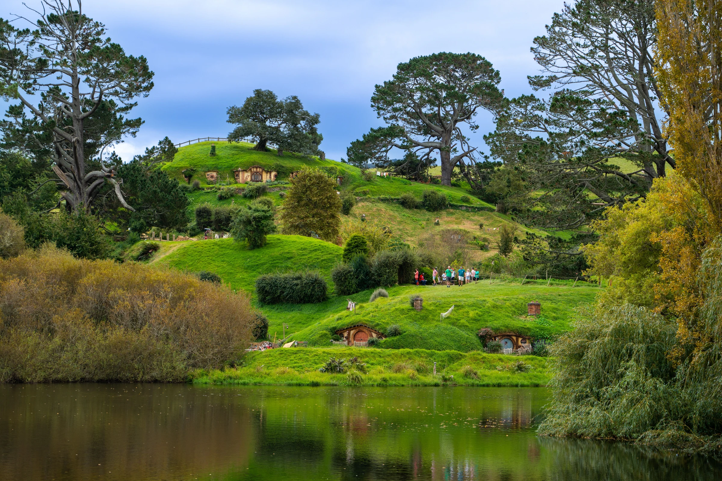 Hobbiton movie-set houses on green hills beside a pond