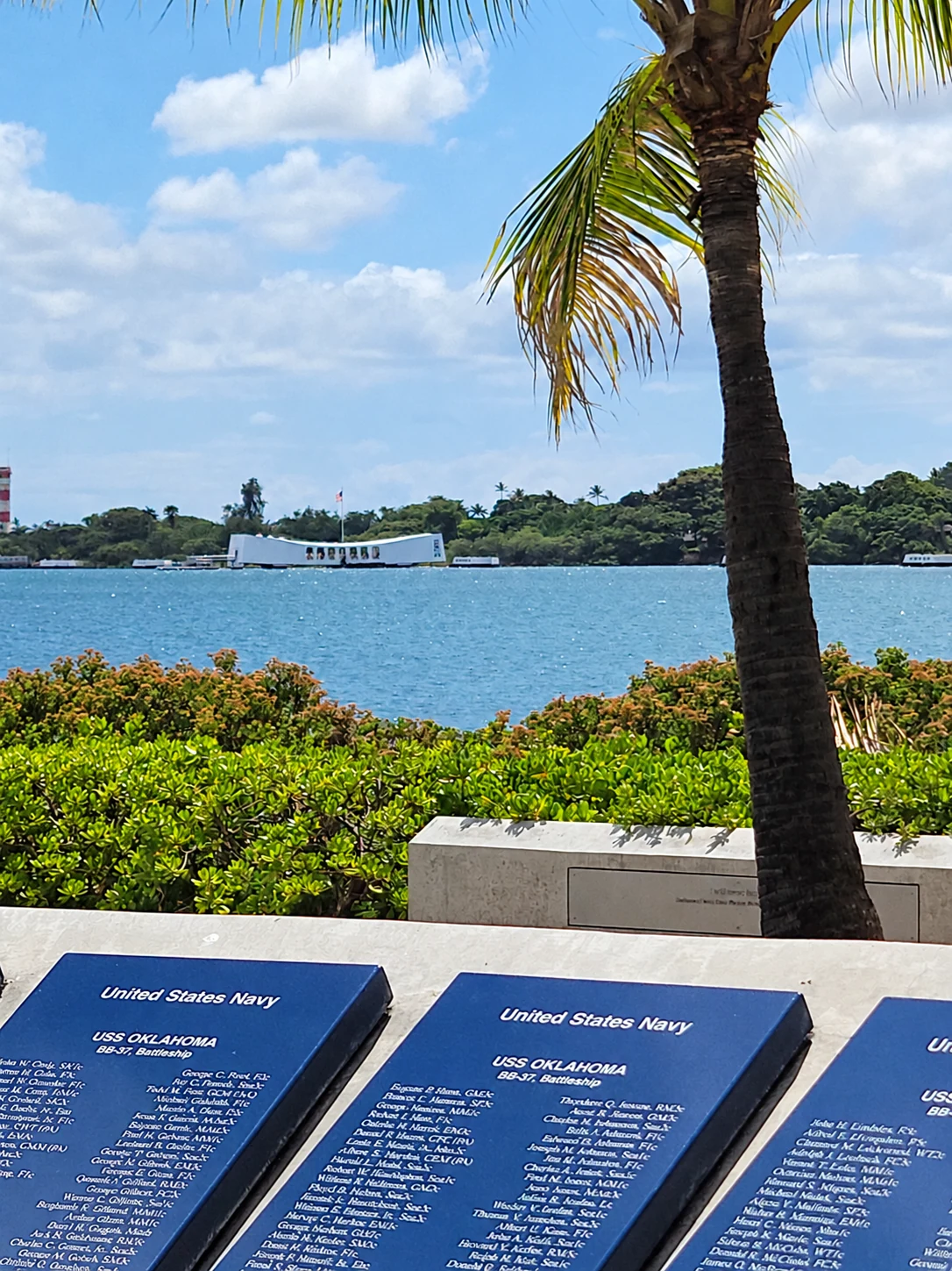 USS Arizona Memorial across Pearl Harbor, framed by palms and memorial plaques