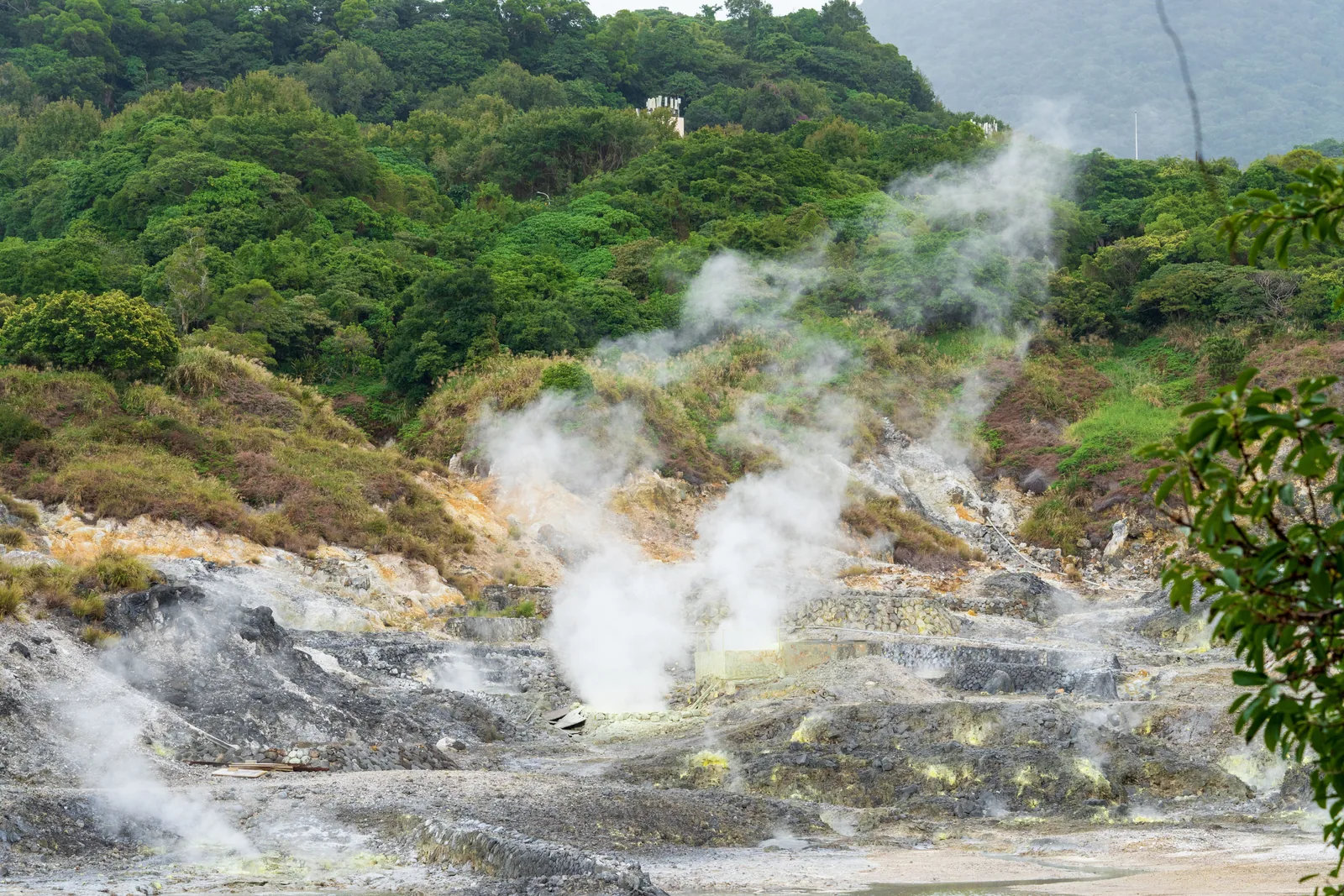 Steam rising from rocky geothermal ground surrounded by green hills