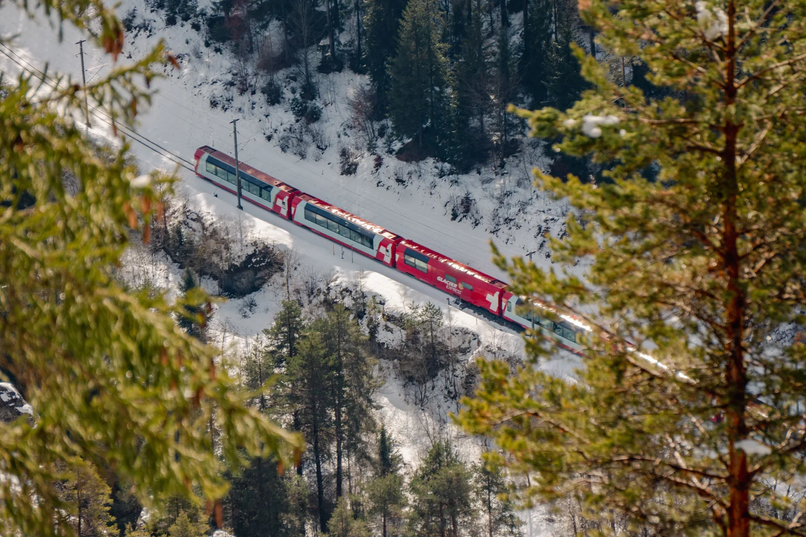 Red Swiss passenger train beside forest and snowy mountains