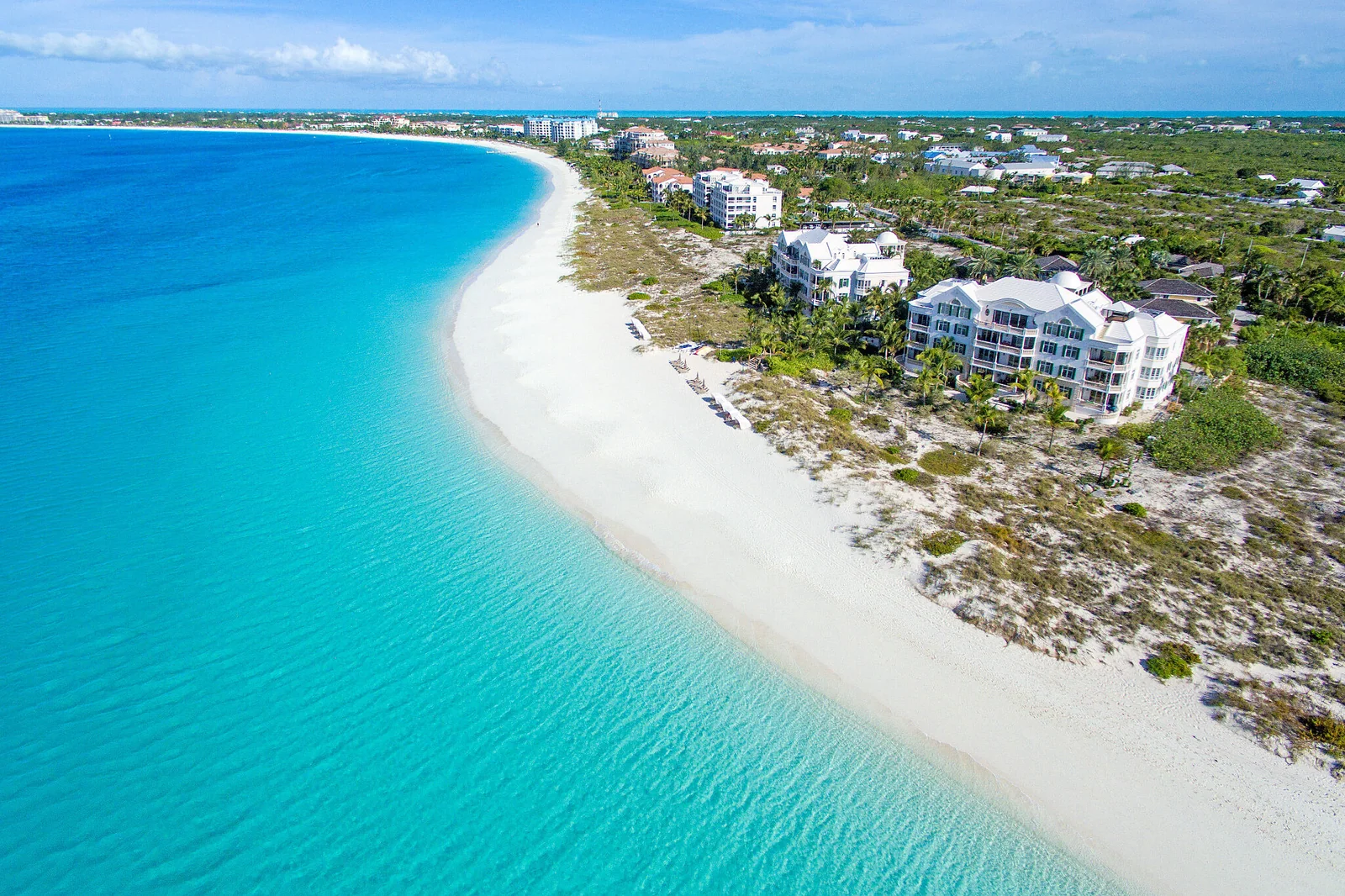 An aerial view of a long curving white-sand beach beside turquoise water and coastal buildings
