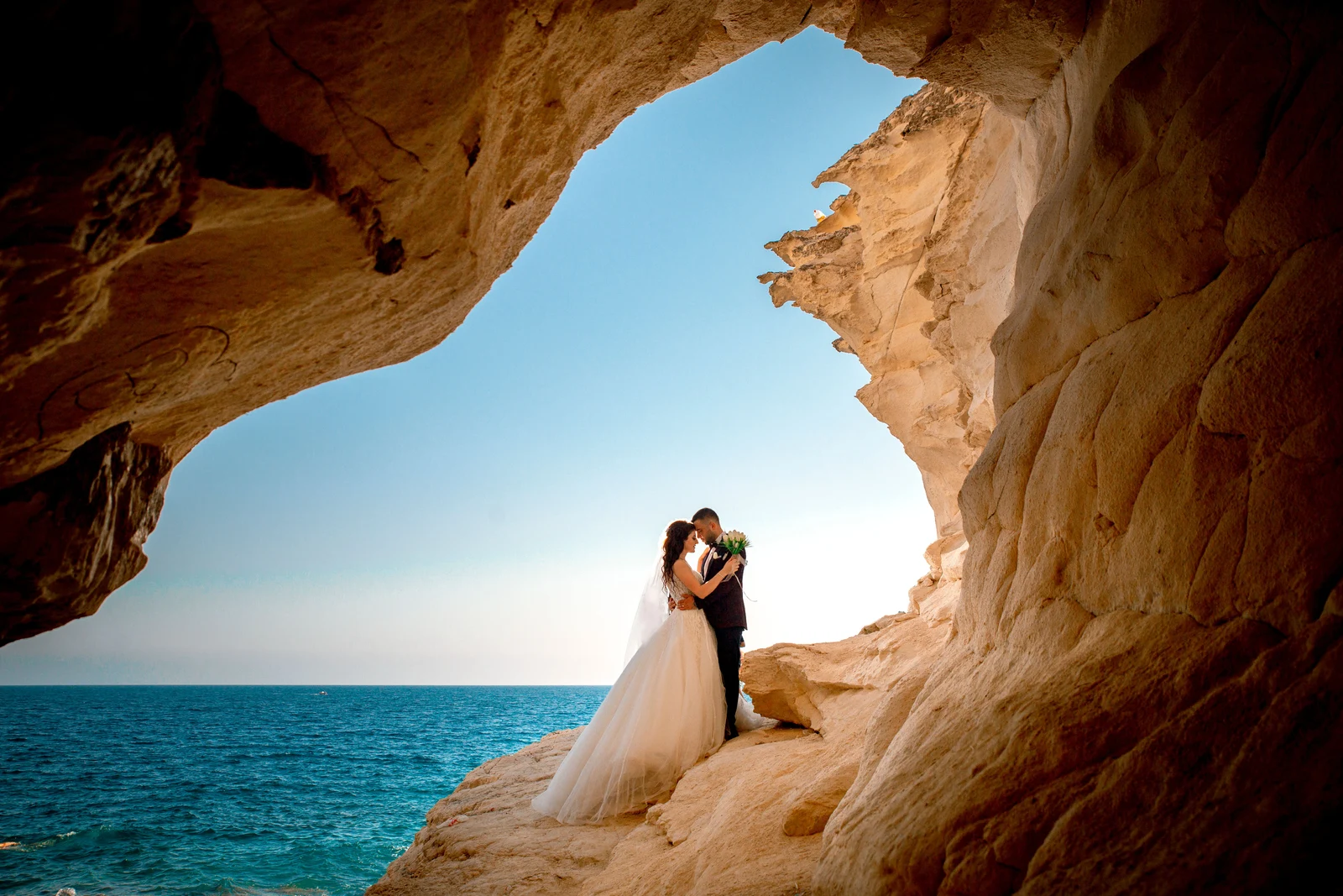 Newlyweds embracing beneath a coastal rock arch overlooking the sea