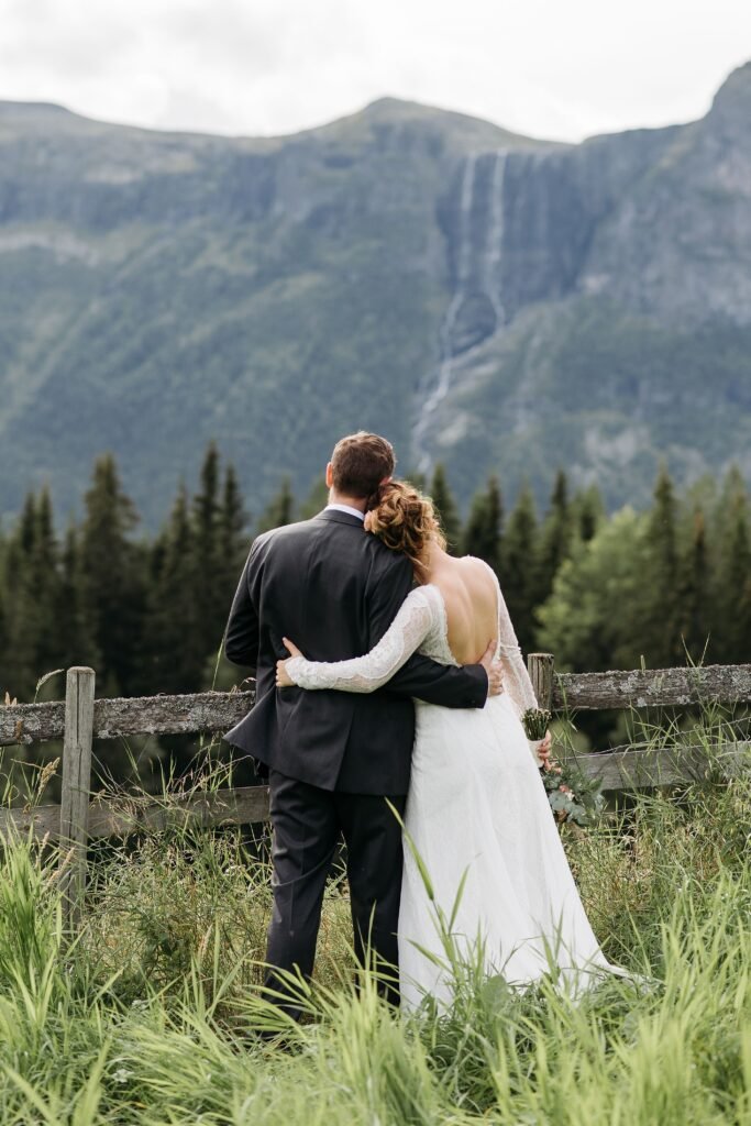 Newlyweds embracing beside a wooden fence with mountains and a waterfall beyond