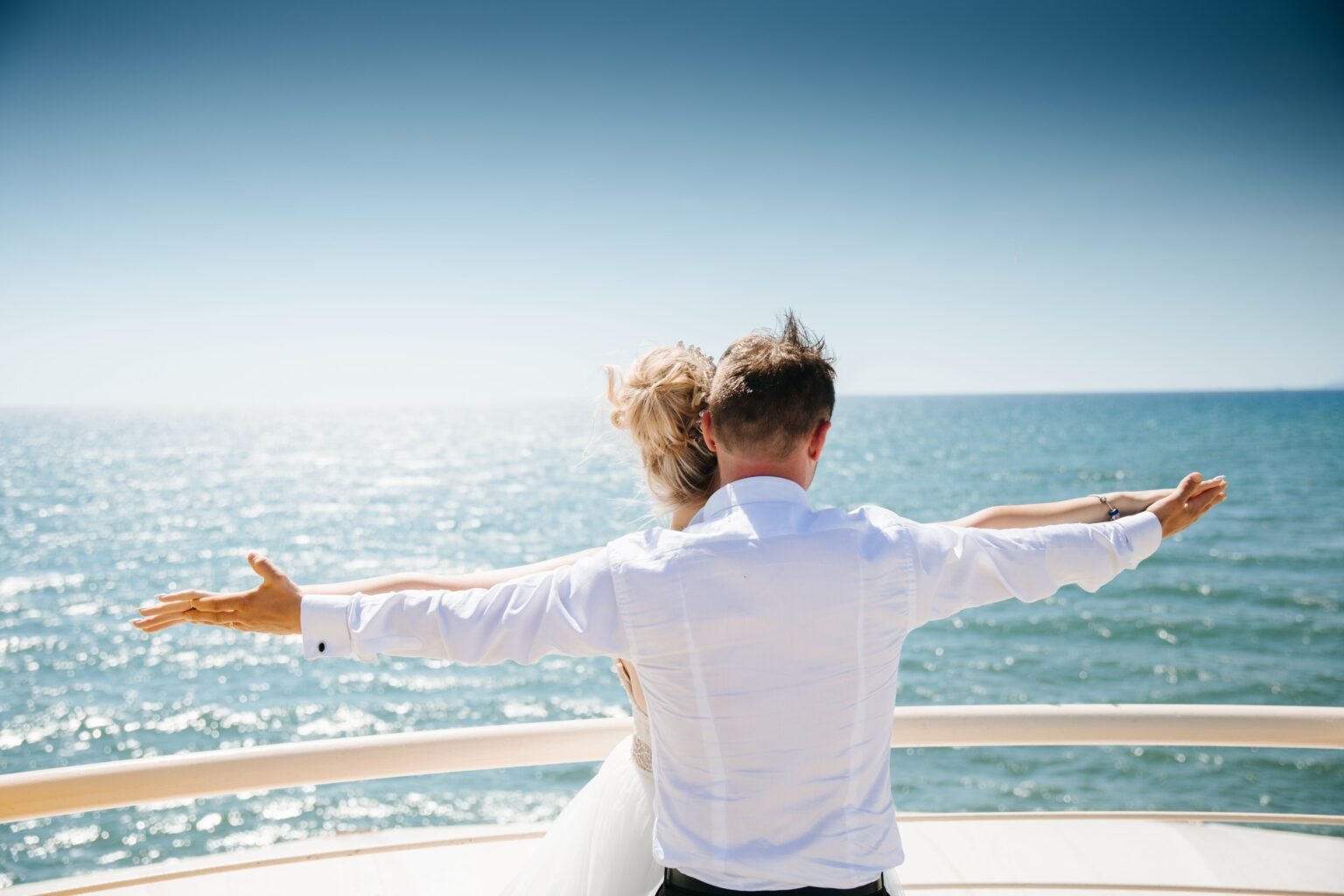A couple standing together at a ship railing with arms outstretched toward the sea